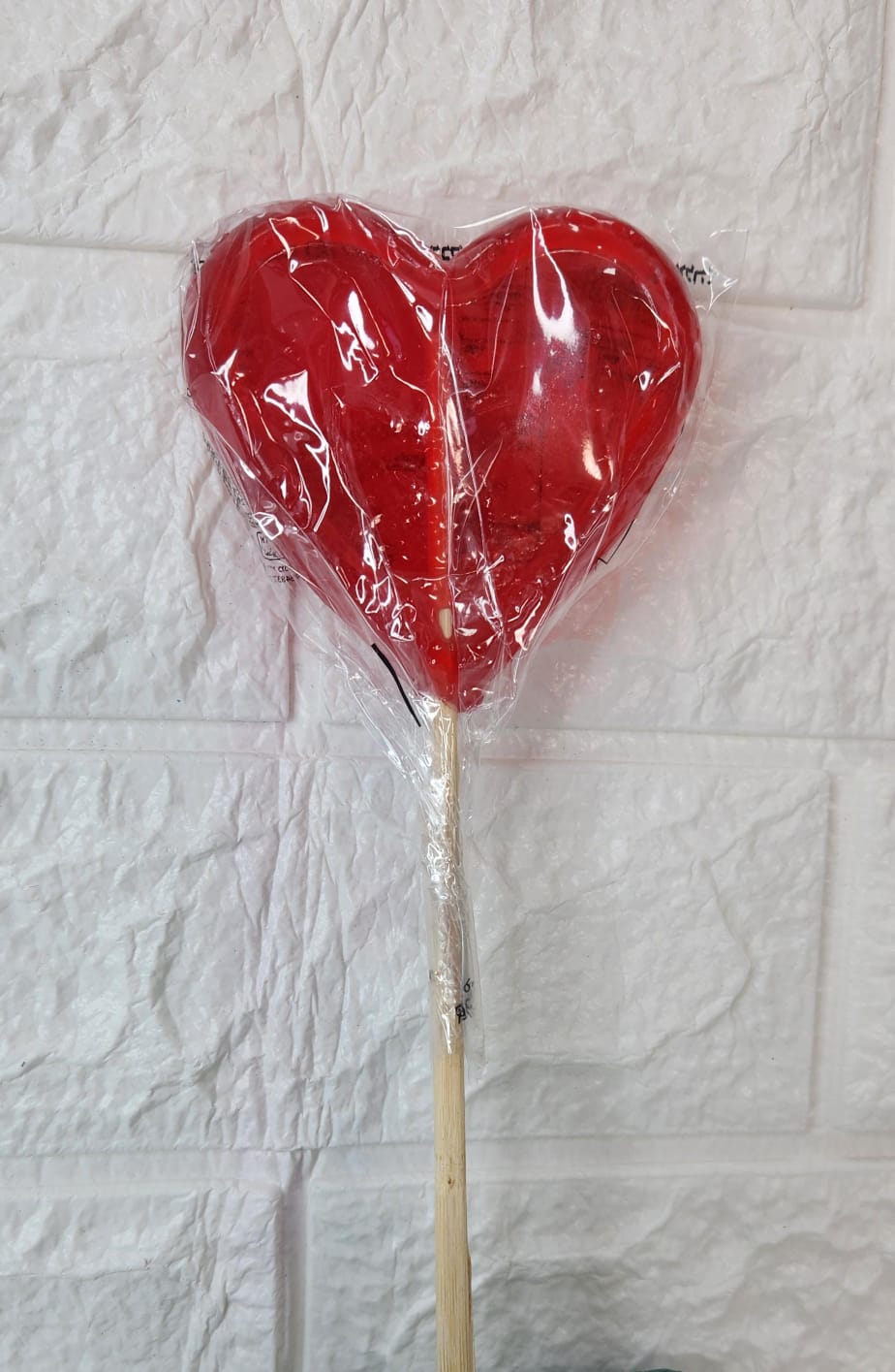 עוגת סוכריות בצורת לב אדומה עטופה בניילון, מחוברת למקל עץ.