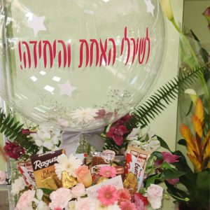 זֵר פרחים גדול בקופסה, מתחת לבלוּן שקוף עם הכיתוב "לשירלי האחת והיחידה!" וממתקים.