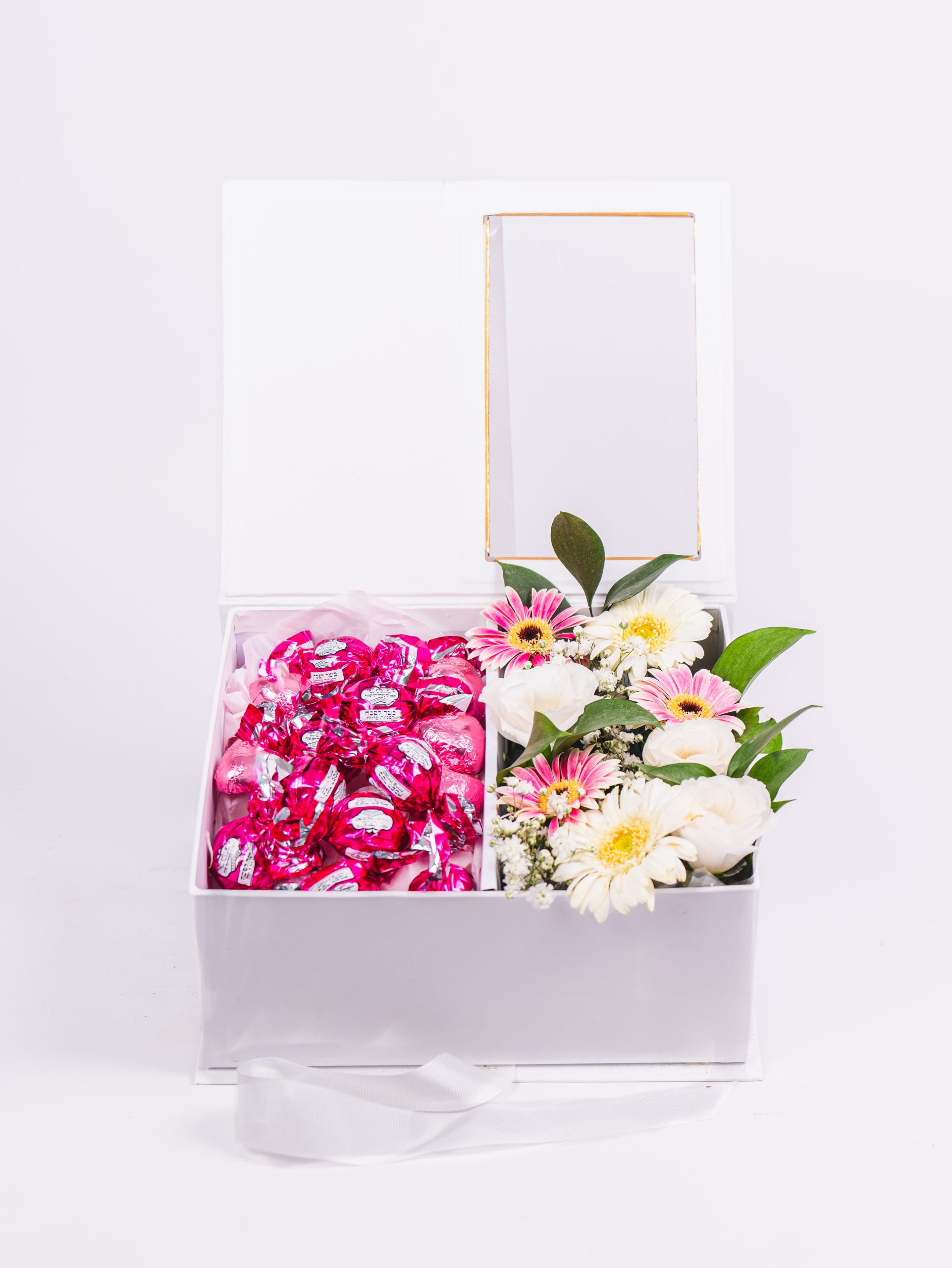 קופסה לבנה פתוחה עם שוקולדים ורודים וזר פרחים לבנים וורודים.