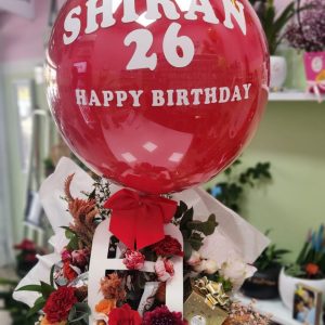 בלון אדום גדול עם הכיתוב "שירן 26 יום הולדת שמח" מעל זר פרחים אדומים, לבנים וכתומים בקופסה.
