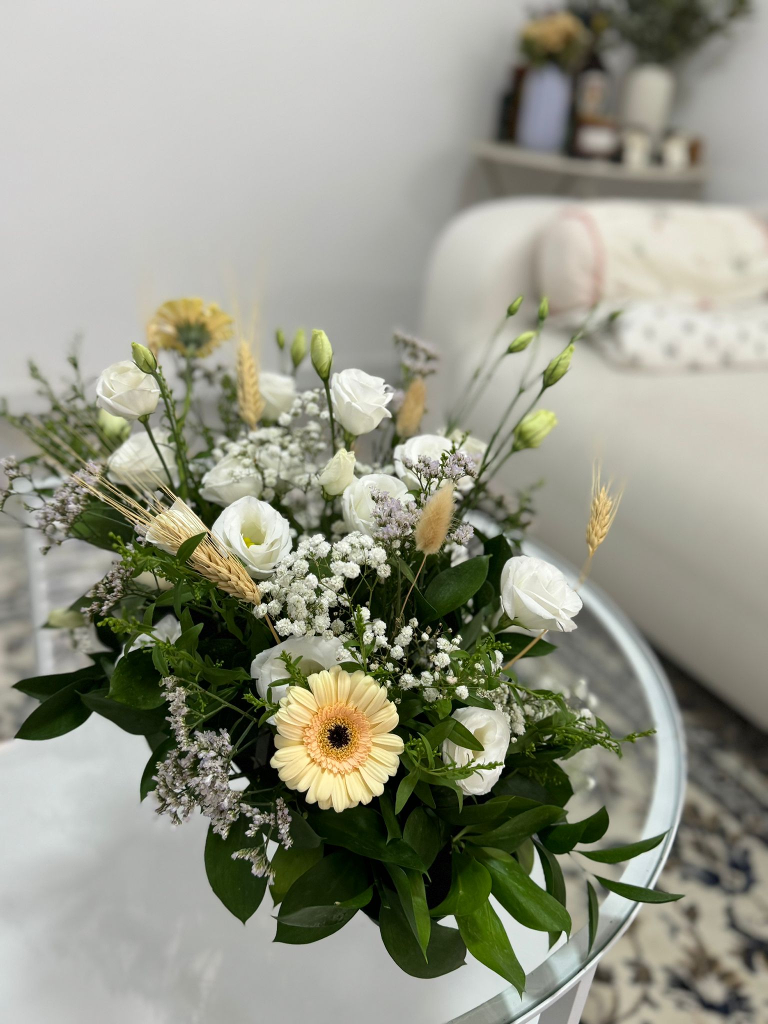 זר פרחים לבנים וצהובים, עם עלי ירק ושיבולים, מונח על שולחן זכוכית עגול.