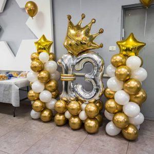 קישוטים ליום הולדת 13: בלונים בצבעי זהב ולבן מסודרים בעמודים, בלון כתר זהב גדול ומספר 13 מבלוני כסף גדולים.