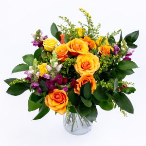 זר פרחים צבעוני באגרטל זכוכית, עם ורדים כתומים וצהובים, פרחים סגולים ועלים ירוקים, על רקע לבן.