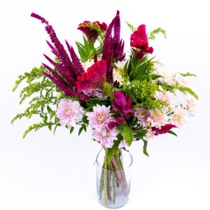 זר פרחים צבעוניים, ורודים, סגולים וירוקים, מונח באגרטל זכוכית על רקע לבן.