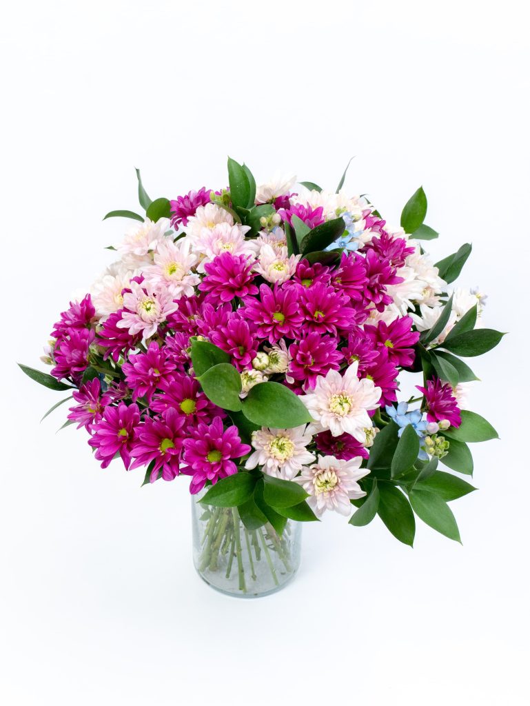 זר פרחים סגולים, לבנים וורודים עם עלים ירוקים, מונח באגרטל זכוכית על רקע לבן.