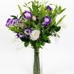 זר פרחים סגולים ולבנים עם עלים ירוקים וכמה פרחים קטנים לבנים, מונח בתוך אגרטל זכוכית שקוף על רקע לבן.