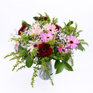 זֵר פרחים גדול וצבעוני, הכולל גרברות בגוונים ורודים, אדומים ולבנים, וענפים ירוקים, עומד בתוך צנצנת זכוכית שק