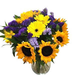 זר פרחים צבעוני באגרטל שקוף, עם חמניות צהובות, גרברות צהובות ופרחים סגולים קטנים.