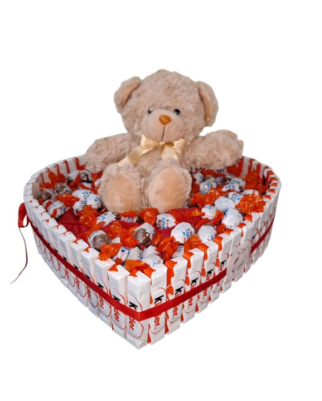 בובת דוב חום יושבת בתוך קופסת לב מלאה בסוכריות קינדר וסוכריות עטופות.