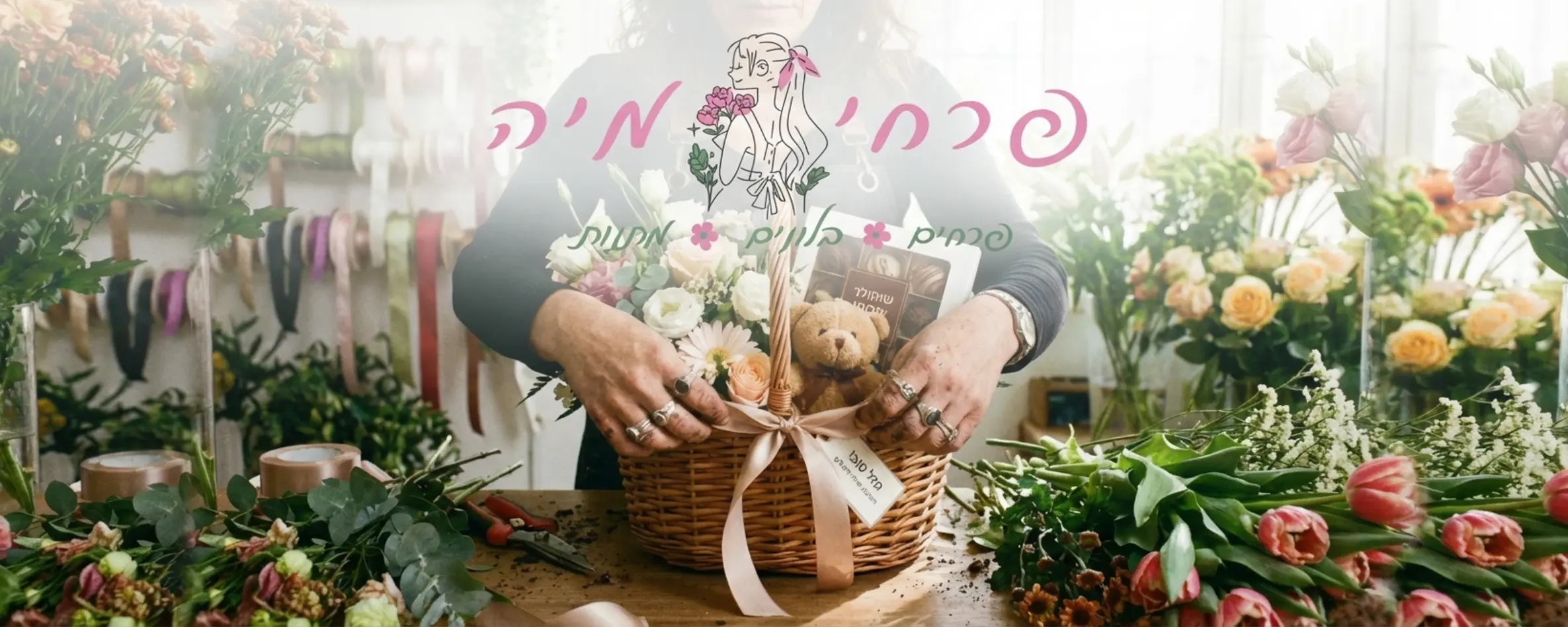 צילום קרוב של שוזרת פרחים מקצועית מסדרת מארז מתנה ליולדת הכולל פרחים טריים, דובי פרווה קטן וקופסת שוקולדים, כשהיא קושרת סרט סאטן תואם מסביב למארז בסביבת עבודה מלאה בפרחים