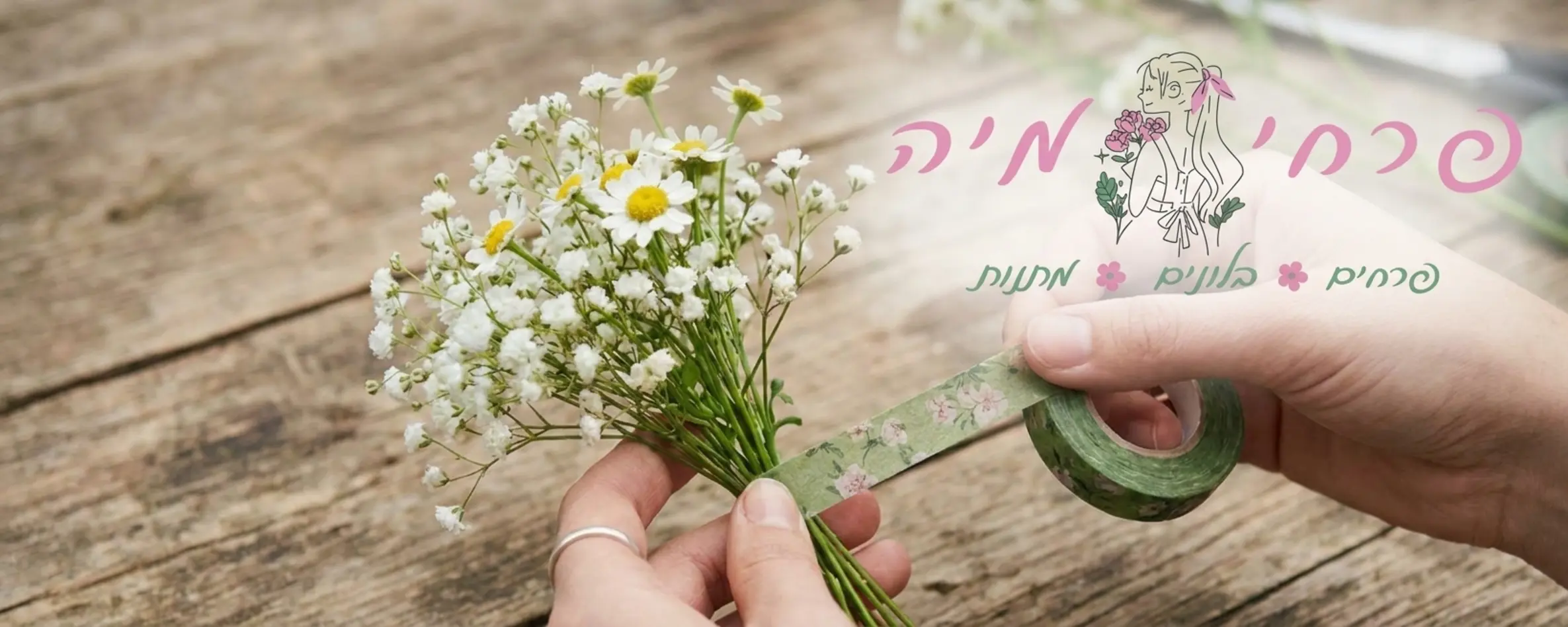 צילום תקריב של ידיים עדינות האוחזות בקבוצה קטנה של פרחי גיבסנית וחרציות לבנות ומלפפות סביב הגבעולים סרט הדבקה פרחוני בצבע ירוק על רקע שולחן עץ טבעי
